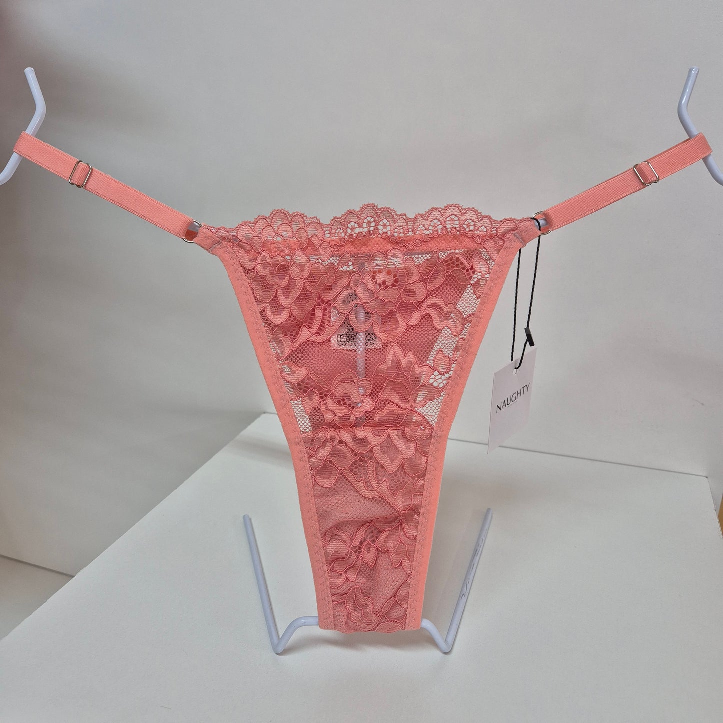 Calcinha string coral em renda macia e floral com alças finas ajustáveis e toque artesanal. Fio duplo em microfibra e plaquinha personalizada naughty na cor prata. 